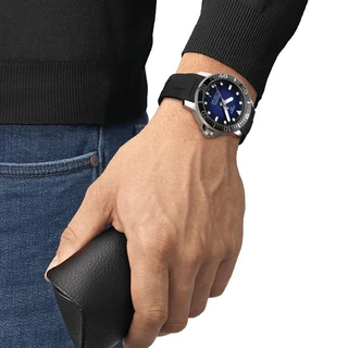 שעון יד לגבר Tissot עם רצועת סיליקון שחורה, חוגה כחולה, מוצג בזווית קרובה על פרק יד.